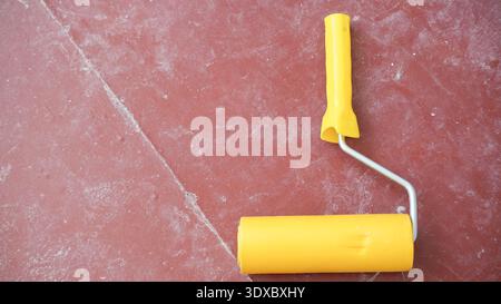 Yellow paint roller lying on dusty red floor surface prepared for painting during home renovation project. Stock Photo