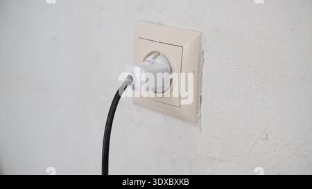 White electrical plug connected to wall socket on textured plaster surface inside renovated room. Stock Photo