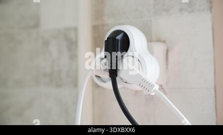 Multiple electrical plugs connected to a wall socket adapter in a tiled kitchen interior. Stock Photo