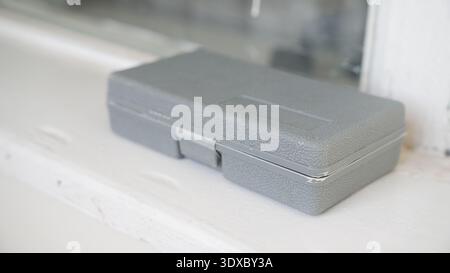 Gray textured plastic tool case placed on white windowsill with soft natural light and minimalist interior background. Stock Photo