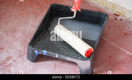 Paint roller resting in black tray filled with white primer on red floor during home renovation process. Stock Photo
