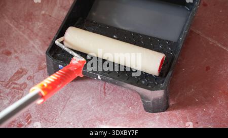 Paint roller with red handle resting in black tray filled with white paint on dusty red floor during renovation. Stock Photo