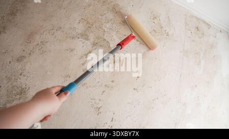 Hand holding extension pole with paint roller applying primer to stained interior wall surface. Stock Photo