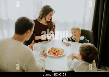 A family sits around a table in their home enjoying breakfast. The parents drink coffee while the children look at a fruit plate. The sun shines throu Stock Photo