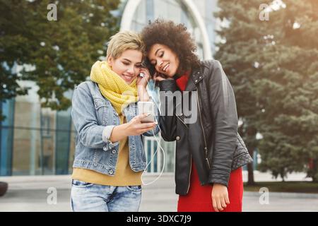 Two Friends Share Music on a Phone in an Outdoor Setting With Trees and Modern Buildings Stock Photo