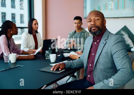 Confident mature businessman in smart casual sitting at meeting table with colleagues in boardroom Stock Photo