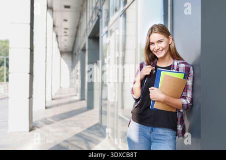 Smiling Student Stands in a Modern Building Holding Books and Folders in a Bright Setting Stock Photo