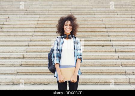Young Person Smiles at Camera While Standing on Steps With Backpack and Headphones in Daytime Stock Photo