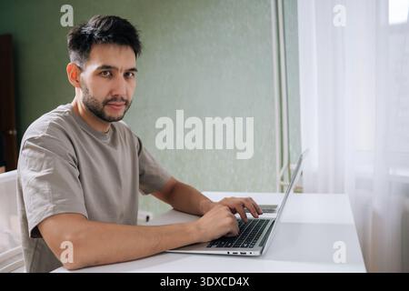 Portrait of bearded programmer male typing on laptop computer looking at camera. Concept of remote work, technology use and freelance productivity Stock Photo