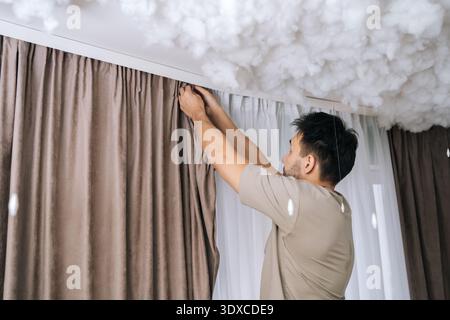 Shooting from below of man hanging curtains on modern curtain rail, installing new window treatments in stylish bedroom, engaging in home improvement Stock Photo