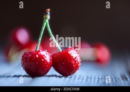 Two ripe cherries with stems on rustic wooden surface, shallow depth of field. Fresh red sweet fruit with water droplets, summer harvest concept Stock Photo