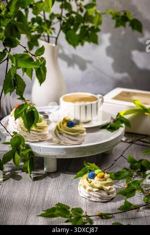 Individual meringue nests with pistachio cream and colorful chocolate eggs arranged on a wooden cake stand beside a cup of coffee, showcasing an elega Stock Photo