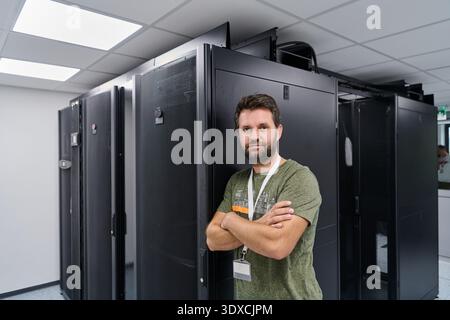 IT technician standing confidently in a data center server room next to black server racks with badge Stock Photo