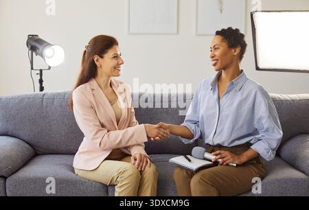 Portrait of young cheerful journalist shaking hands greeting her guest sitting in studio. Stock Photo