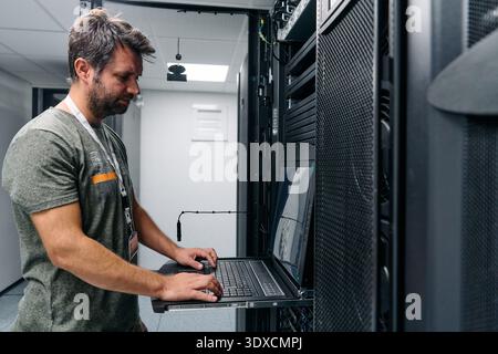 It technician configuring servers on a rack-mounted laptop in a modern data center server room Stock Photo
