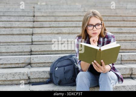 Student Reads a Book While Sitting on Stone Steps With a Backpack Beside Her Stock Photo
