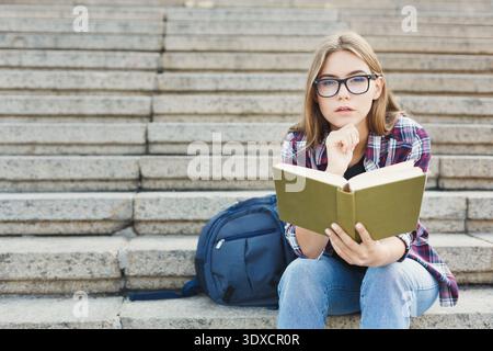 Young Person Reads a Book While Sitting on Steps With a Backpack Nearby in a Public Area Stock Photo