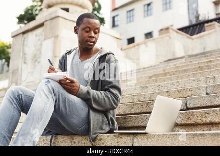 Man Writes in Notebook While Sitting on Steps Near Laptop During Daytime Stock Photo