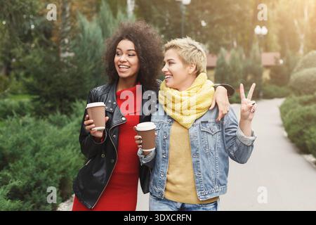 Two Friends Walk in a Park and Enjoy Coffee While Talking and Having Fun Together in Autumn Stock Photo