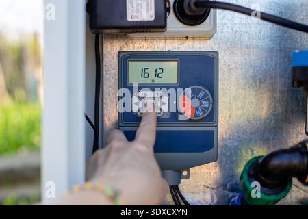 A hand operates a digital irrigation timer on a control panel in a garden. smart farming, automated watering, modern technology. Stock Photo