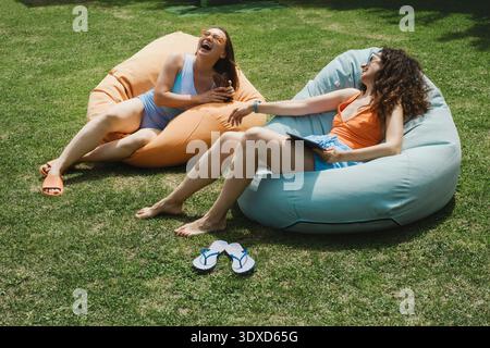 Two female friends relax on beanbag chairs on a green lawn using a tablet and chatting. They enjoy a sunny summer day in the backyard during vacation. Stock Photo