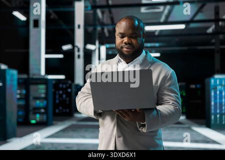 Computer scientist in data center updating systems using laptop, protecting equipment from damaging software errors. Server hub employee using notebook, looking for bugs causing gear to slow down Stock Photo