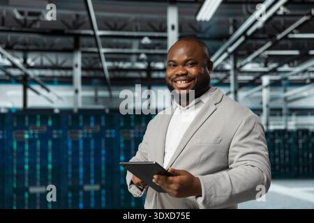 Cheerful engineer in data center updating systems using tablet, protecting equipment from damaging software errors. Happy server farm worker using device, looking for bugs causing gear to slow down Stock Photo