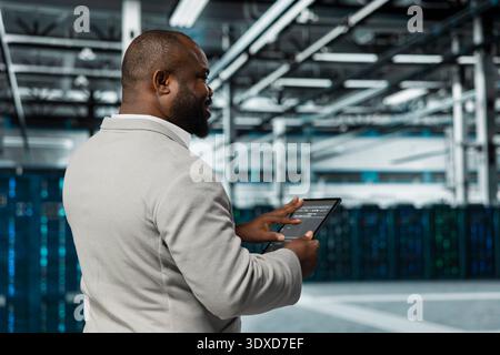 System administrator walking in data center running lines of code on tablet, troubleshooting rackmounts. Employee using programming scripts on device in server farm, optimizing performance Stock Photo