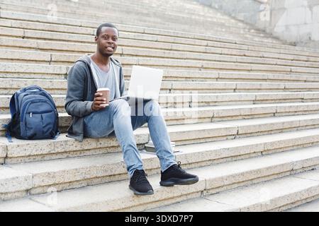 Young Man Sits on Stairs Using Laptop and Drinking Coffee in Urban Setting During Daytime Stock Photo