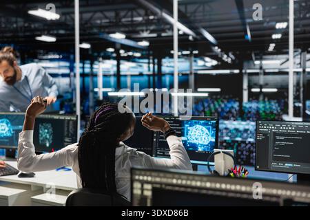 Exhausted data center female programmer stretching to ease back tension while using artificial intelligence. African american woman in server farm relieving pain after long hours at computer Stock Photo