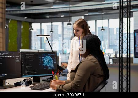 Senior engineer showing junior dev how to adjust deep learning algorithms parameters to automate tasks. Tech company team leader teaching intern to use AI driven insights for code optimization Stock Photo