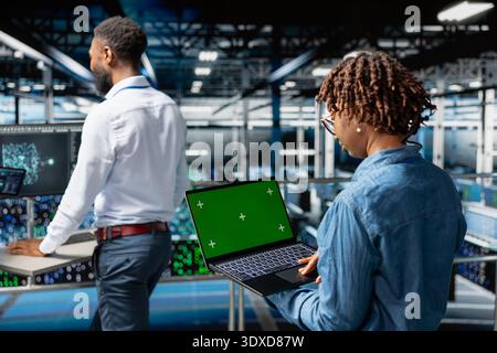 Server farm black engineer holding isolated screen on tablet to review diagnostics for performance stability in data center. Female employee on elevated industrial platform solving tasks. Stock Photo