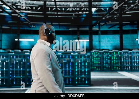 Engineer working in data center, listening music and overseeing infrastructure mainframes. Employee wearing audio headphones walking in server farm delivering web content to online customers Stock Photo