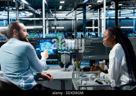 Data center manager and intern monitoring AI applications, adjusting machine learning parameters. Server hub supervisor helping trainee enhance artificial intelligence software performance Stock Photo