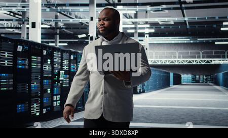 Data center system administrator tasked with reviewing code for AI automation scripts. African american man walking in server hub running diagnostics on equipment rigs, camera A Stock Photo
