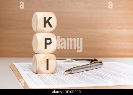 Front view of three wooden toy blocks spelling the acronym KPI stacked vertically on a business desk with a pen and report. Concept of key performance Stock Photo