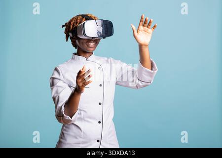 Black woman chef interacts with virtual reality interface, exploring digital cooking techniques. Female cook uses AR technology to practice culinary skills and prepare for upcoming competitions. Stock Photo