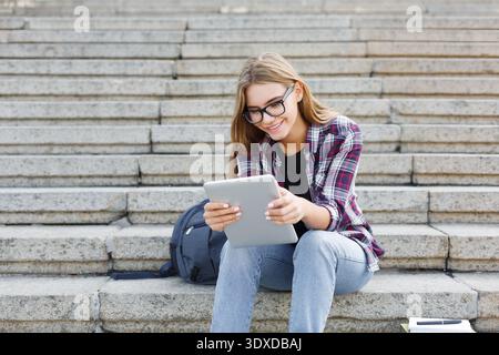Student Enjoys Using Tablet While Sitting on Steps in Urban Area During Sunny Day Stock Photo