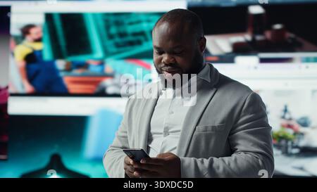 Portrait of cheerful man browsing social media feed on smartphone in front of digital video wall. Happy person using mobile phone, floating icons highlight interactive engagement online, camera A Stock Photo