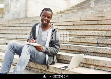 Student Works on Notes in Outdoor Setting Using Laptop and Notebook on Stone Steps Stock Photo