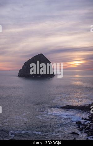 Golden sun dips behind Haystack Rock at Cape Kiwanda on the Pacific coast of Oregon, USA. Lonely sea stack stands bold against colorful sky. Waves gen Stock Photo