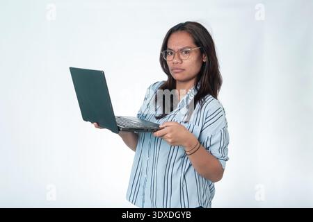 Portrait of a young Indonesian woman in glasses holding a laptop with a focused, serious expression. Concept for work, education, and digital life. Stock Photo