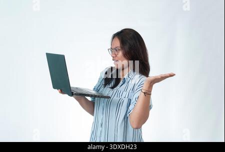 Portrait of a surprised Indonesian woman holding a laptop with a questioning gesture. Concept for tech issues, online error, and remote work stress. Stock Photo