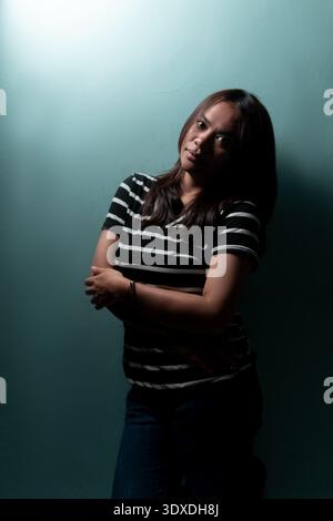 A confident Indonesian woman posing with crossed arms. Dramatic side lighting against a blue wall for moody lifestyle and artistic portrait concepts. Stock Photo