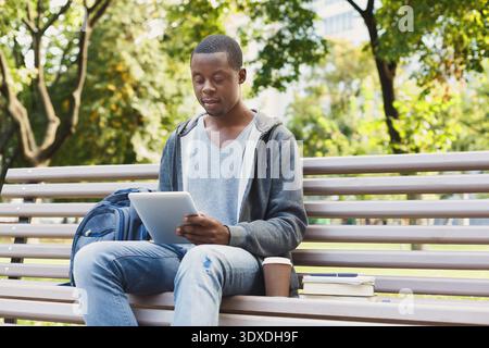 Young Man Sits on Bench in Park Using Tablet for Study and Leisure Activities During Daytime Hours Stock Photo