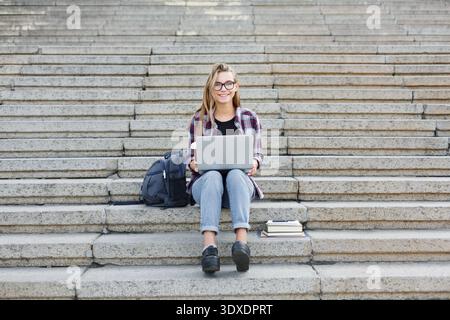 College Student Studies on Stairs With Laptop and Books in a Busy Campus Area During the Day Stock Photo
