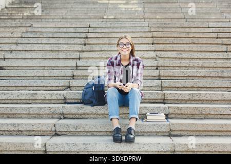 Young Person Sits on Steps With Books and Backpack While Using a Phone Stock Photo