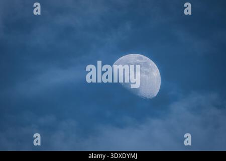 Close-up of a gibbous moon visible during the day, partially covered by soft wispy clouds in a blue sky. Stock Photo