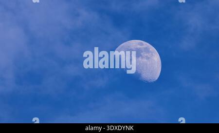 Close-up of a gibbous moon visible during the day, partially covered by soft wispy clouds in a blue sky. Stock Photo