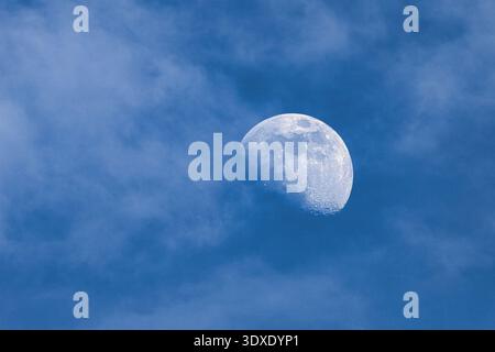 Close-up of a gibbous moon visible during the day, partially covered by soft wispy clouds in a blue sky. Stock Photo